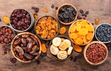 Verschiedene Trockenfrüchte in Schalen auf einem Holztisch aus der Vogelperspektive: Datteln, Feigen, Aprikosen, Pflaumen und verschiedene Rosinen.