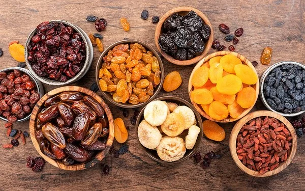 Verschiedene Trockenfrüchte in Schalen auf einem Holztisch aus der Vogelperspektive: Datteln, Feigen, Aprikosen, Pflaumen und verschiedene Rosinen.