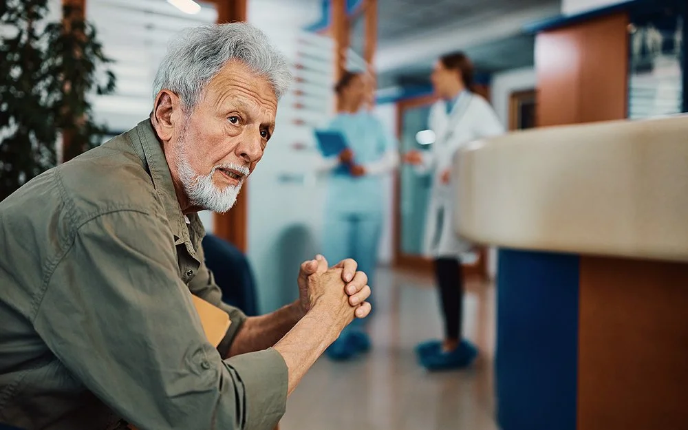 Ein älterer Mann sitzt mit besorgtem Blick im Wartebereich einer Arztpraxis oder eines Krankenhauses.