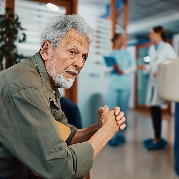 Ein älterer Mann sitzt mit besorgtem Blick im Wartebereich einer Arztpraxis oder eines Krankenhauses.