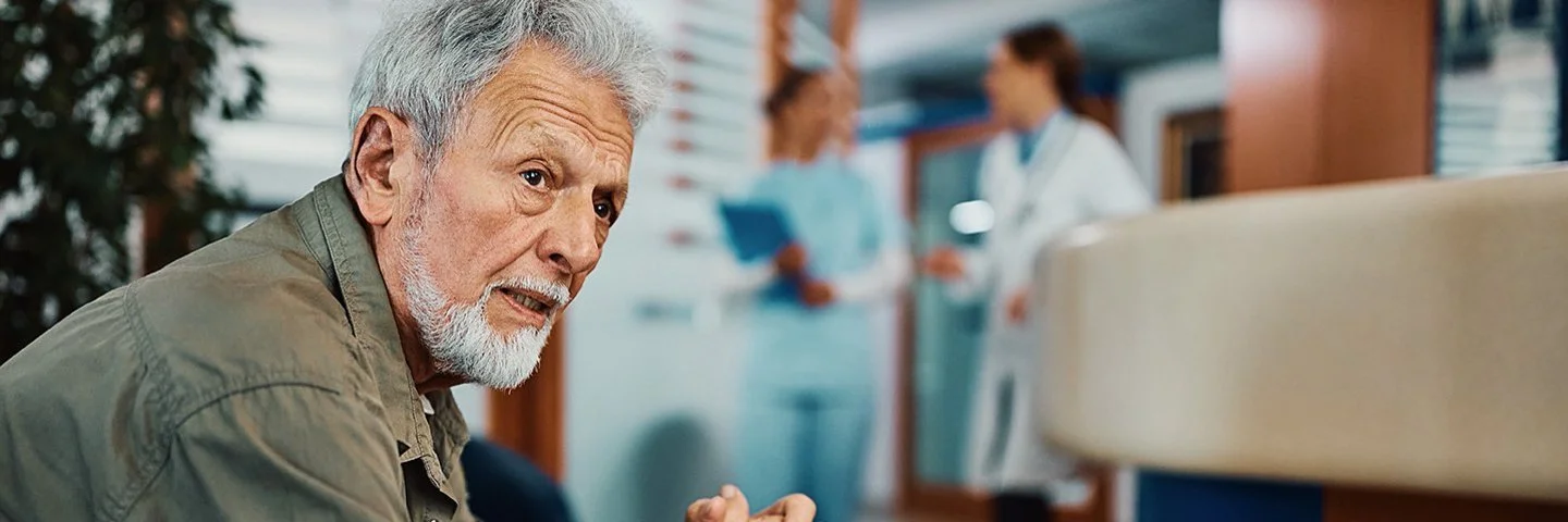 Ein älterer Mann sitzt mit besorgtem Blick im Wartebereich einer Arztpraxis oder eines Krankenhauses.