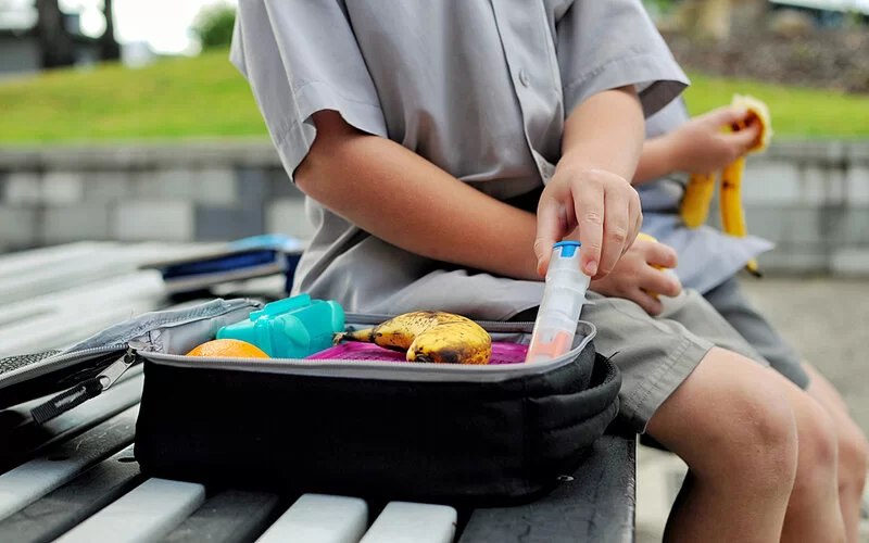 Auf einer Bank sitzt ein Kind und macht Mittagspause. Aus seiner Lunchbox holt es einen Epipen.