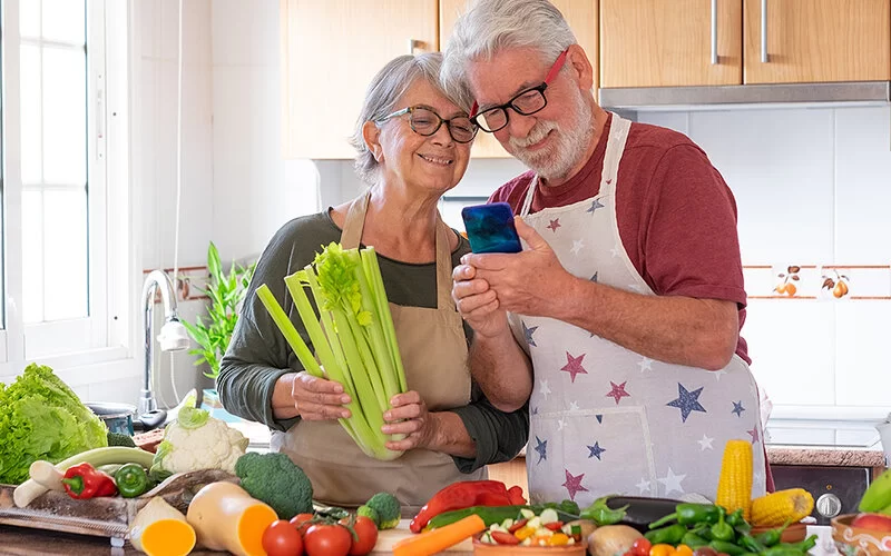 Gesund essen für den Blutdruck: Eine ältere Frau und ein älterer Mann stehen in der Küche, vor ihnen liegen viele gesunde Lebensmittel, auf ihrem Handy schauen sie das Rezept an.