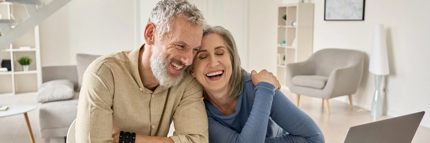 Glückliches, reifes Ehepaar im besten Alter, das lachend und eng verbunden am Tisch zu Hause mit einem Laptop sitzt.