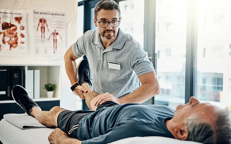 Ein Physiotherapeut macht mit einem liegenden älteren Patienten Beinübungen, um die Muskelkraft bei Myositis zu verbessern.