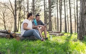 Zwei Personen sitzen auf gestapelten Baumstämmen in einem lichten Wald. Die Person links trägt ein weißes T-Shirt und graue Leggings, hält eine Wasserflasche und blickt zur anderen Person. Die Person rechts trägt ein dunkles T-Shirt und kurze Hose, hält ein Smartphone und sitzt entspannt. Sonnenlicht fällt durch die Bäume auf den grünen Waldboden.