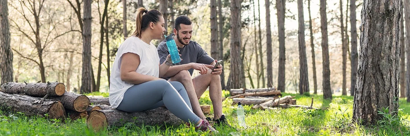 Zwei Personen sitzen auf gestapelten Baumstämmen in einem lichten Wald. Die Person links trägt ein weißes T-Shirt und graue Leggings, hält eine Wasserflasche und blickt zur anderen Person. Die Person rechts trägt ein dunkles T-Shirt und kurze Hose, hält ein Smartphone und sitzt entspannt. Sonnenlicht fällt durch die Bäume auf den grünen Waldboden.