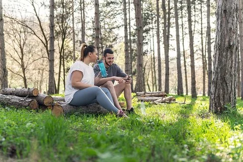 Zwei Personen sitzen auf gestapelten Baumstämmen in einem lichten Wald. Die Person links trägt ein weißes T-Shirt und graue Leggings, hält eine Wasserflasche und blickt zur anderen Person. Die Person rechts trägt ein dunkles T-Shirt und kurze Hose, hält ein Smartphone und sitzt entspannt. Sonnenlicht fällt durch die Bäume auf den grünen Waldboden.