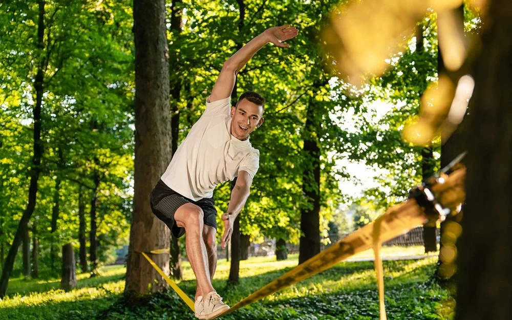 Ein Mann in kurzer Sommerkleidung in einem Park. Er balanciert auf einer Slackline, die zwischen zwei Bäumen gespannt ist.