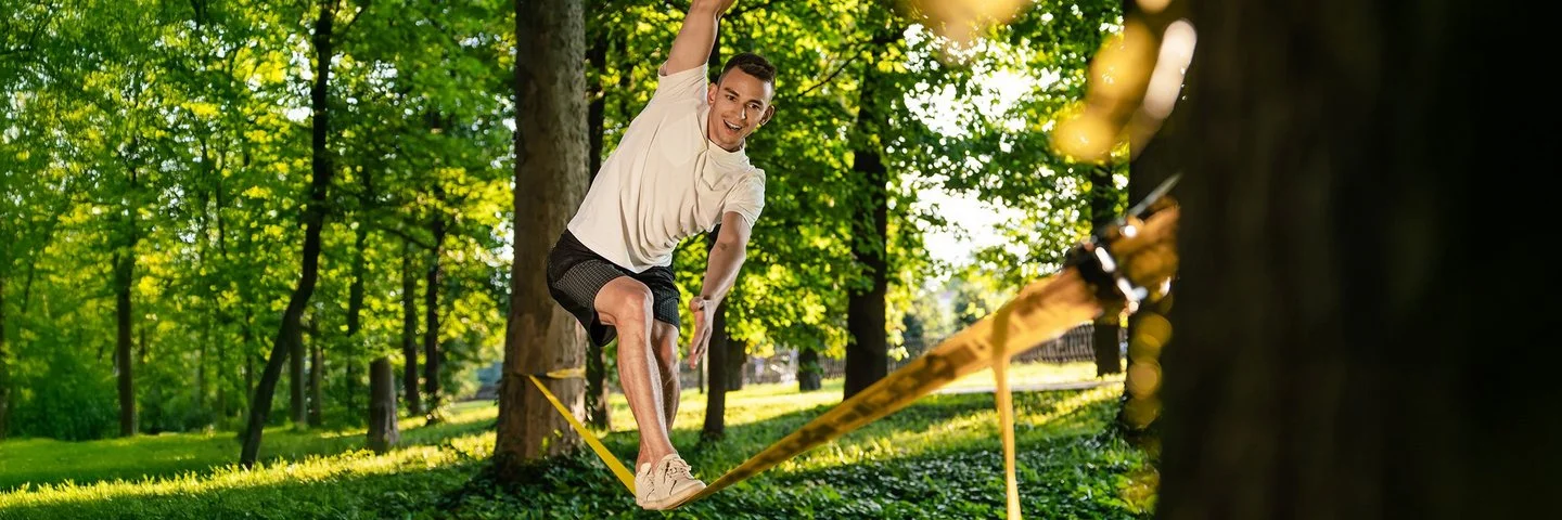 Ein Mann in kurzer Sommerkleidung in einem Park. Er balanciert auf einer Slackline, die zwischen zwei Bäumen gespannt ist.