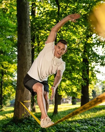 Ein Mann in kurzer Sommerkleidung in einem Park. Er balanciert auf einer Slackline, die zwischen zwei Bäumen gespannt ist.