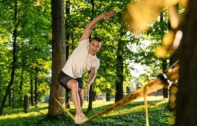 Ein Mann in kurzer Sommerkleidung in einem Park. Er balanciert auf einer Slackline, die zwischen zwei Bäumen gespannt ist.