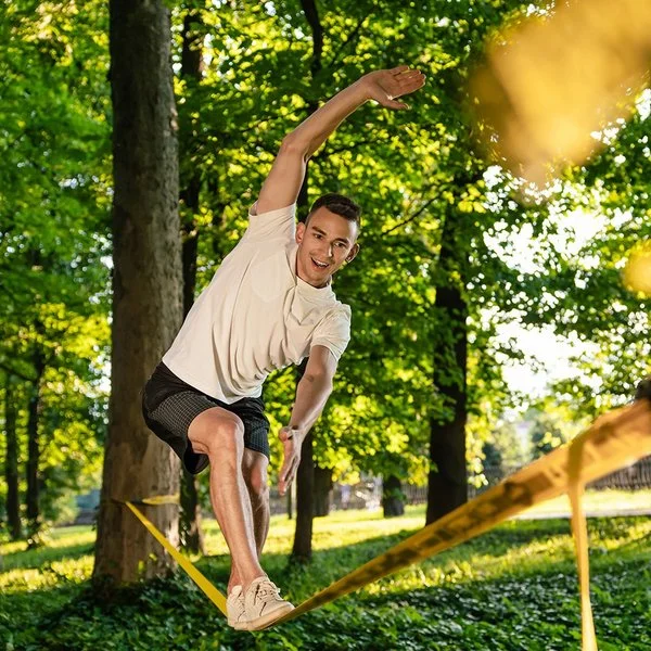 Ein Mann in kurzer Sommerkleidung in einem Park. Er balanciert auf einer Slackline, die zwischen zwei Bäumen gespannt ist.