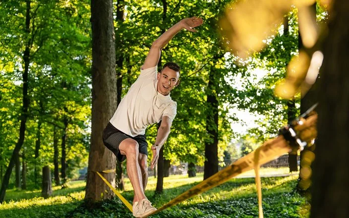 Ein Mann in kurzer Sommerkleidung in einem Park. Er balanciert auf einer Slackline, die zwischen zwei Bäumen gespannt ist.