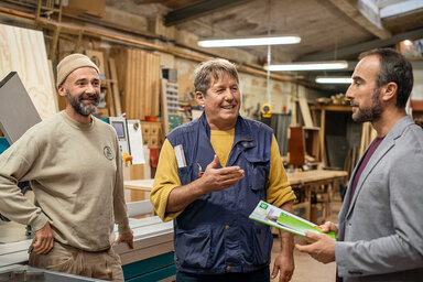 Drei Männer unterhalten sich in einer Werkstatt. Einer trägt ein Jackett und hält Unterlagen, die anderen tragen Arbeitskleidung. Die AOK berät Arbeitgeber.