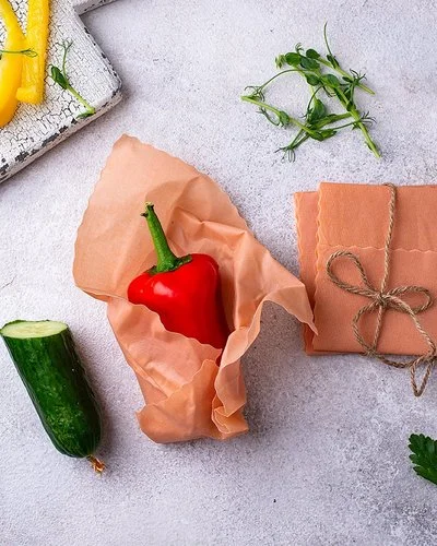 Ein Bienenwachstuch umhüllt eine rote Paprika auf einer hellen Arbeitsfläche. Daneben liegen weitere Tücher und Gemüse.