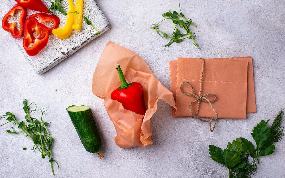 Ein Bienenwachstuch umhüllt eine rote Paprika auf einer hellen Arbeitsfläche. Daneben liegen weitere Tücher und Gemüse.