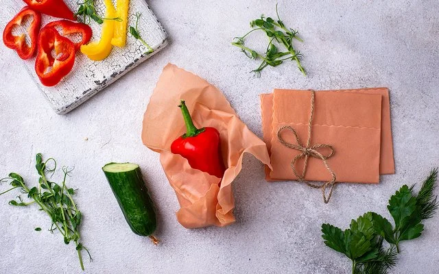 Ein Bienenwachstuch umhüllt eine rote Paprika auf einer hellen Arbeitsfläche. Daneben liegen weitere Tücher und Gemüse.