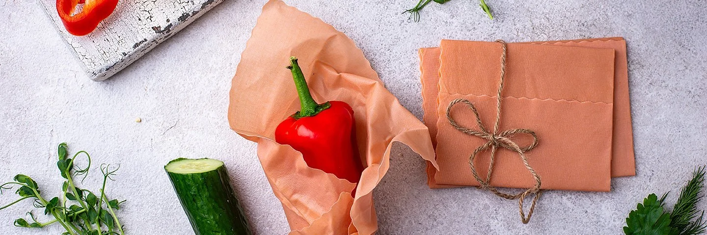 Ein Bienenwachstuch umhüllt eine rote Paprika auf einer hellen Arbeitsfläche. Daneben liegen weitere Tücher und Gemüse.