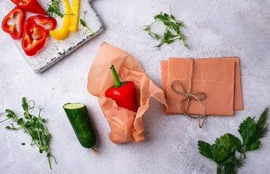 Ein Bienenwachstuch umhüllt eine rote Paprika auf einer hellen Arbeitsfläche. Daneben liegen weitere Tücher und Gemüse.