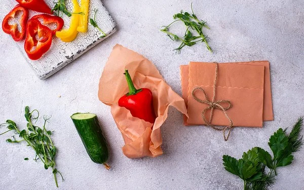 Ein Bienenwachstuch umhüllt eine rote Paprika auf einer hellen Arbeitsfläche. Daneben liegen weitere Tücher und Gemüse.