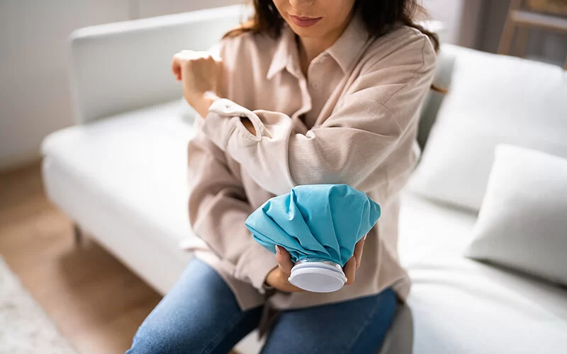 Eine junge Frau sitzt auf der Couch und kühlt ihren blauen Fleck am linken Ellenbogen mit einem Kühlpack.