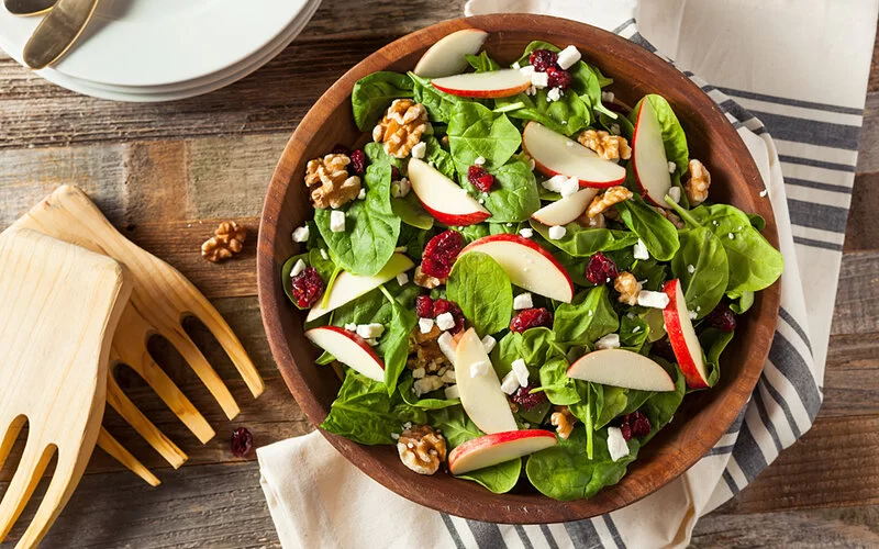 Eine Holzschüssel mit Spinat-Äpfel-Walnuss-Salat auf einem Holztisch.