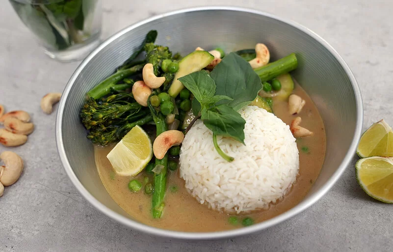 Grünes Gemüsecurry mit Zucchini nach einem Rezept von Felicitas Then, angerichtet auf einem hellgrauen Teller, garniert mit Thai-Basilikum und gerösteten Cashewkernen.