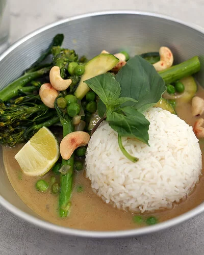 Grünes Gemüsecurry mit Zucchini nach einem Rezept von Felicitas Then, angerichtet auf einem hellgrauen Teller, garniert mit Thai-Basilikum und gerösteten Cashewkernen.