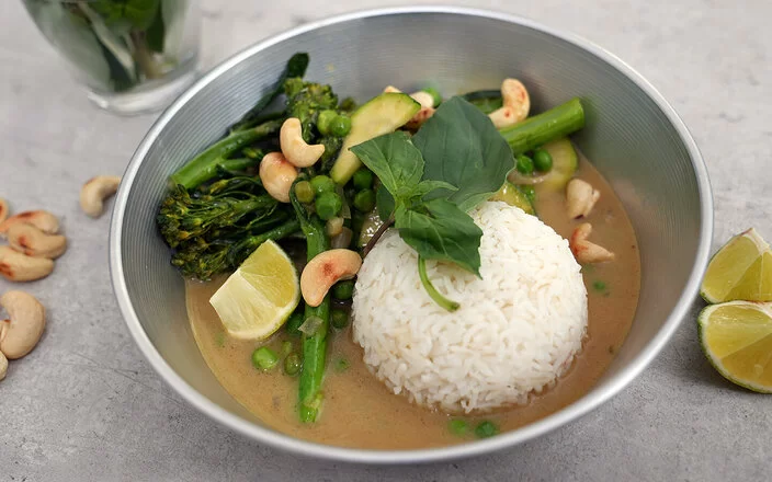 Grünes Gemüsecurry mit Zucchini nach einem Rezept von Felicitas Then, angerichtet auf einem hellgrauen Teller, garniert mit Thai-Basilikum und gerösteten Cashewkernen.