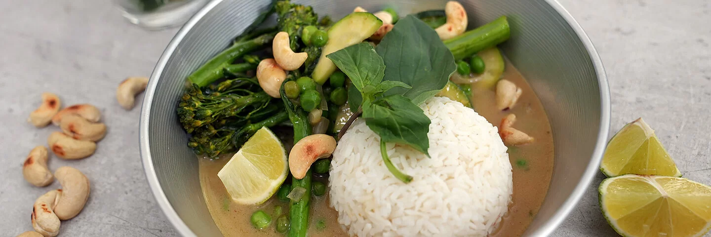Grünes Gemüsecurry mit Zucchini nach einem Rezept von Felicitas Then, angerichtet auf einem hellgrauen Teller, garniert mit Thai-Basilikum und gerösteten Cashewkernen.
