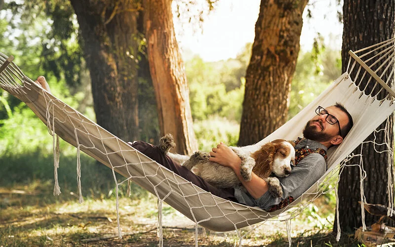 Ein Mann liegt mit seinem Hund auf dem Bauch und mit geschlossenen Augen in einer Hängematte im Freien.