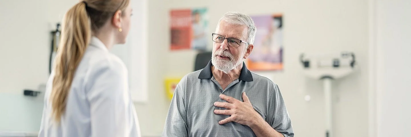 Eine älterer Mann mit Brille und Vollbart spricht beim Check-up mit seiner Ärztin über seine Herzbeschwerden.