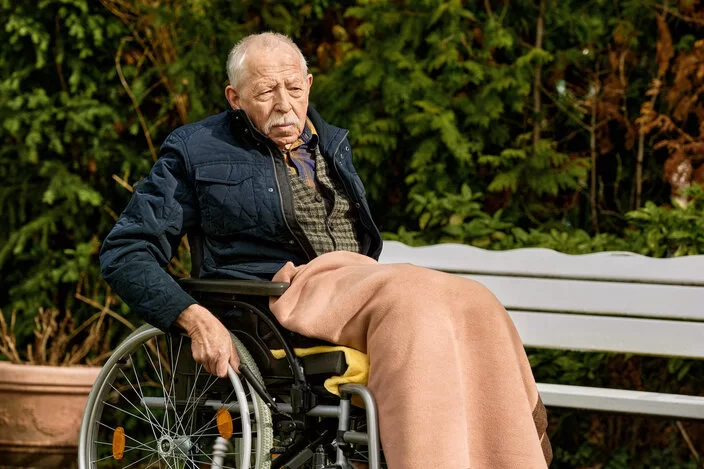 Ein älterer Mann sitzt in einem Rollstuhl und fährt damit durch einen Park.
