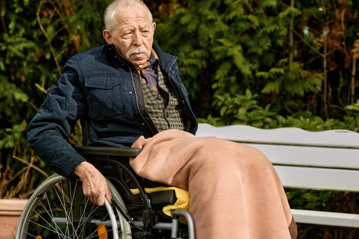 Ein älterer Mann sitzt in einem Rollstuhl und fährt damit durch einen Park.