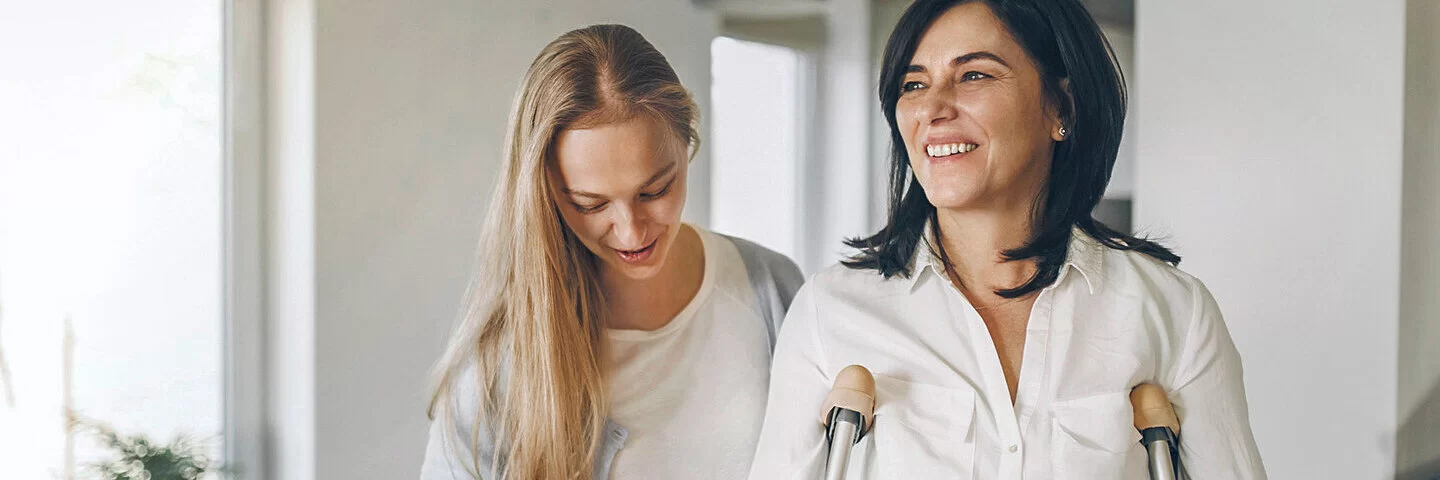 Zwei Frauen in einer Wohnung; eine der Frauen benötigt Gehhilfen, die andere Frau stützt sie.