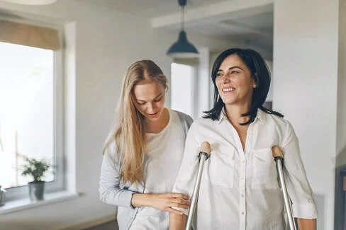 Zwei Frauen in einer Wohnung; eine der Frauen benötigt Gehhilfen, die andere Frau stützt sie.