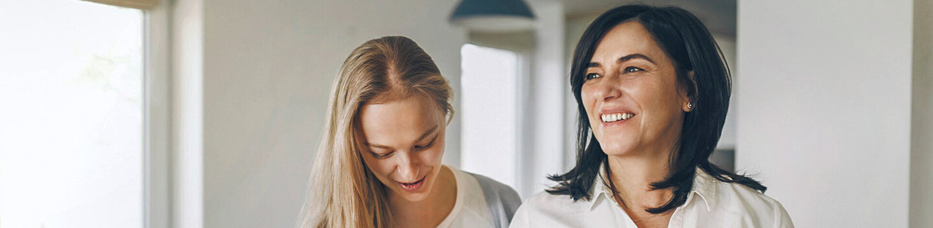 Zwei Frauen in einer Wohnung; eine der Frauen benötigt Gehhilfen, die andere Frau stützt sie.