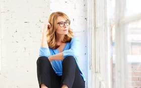 Nachdenklich aussehende mittelalte Frau mit großer Brille sitzt vor einem Fenster und schaut hinaus.