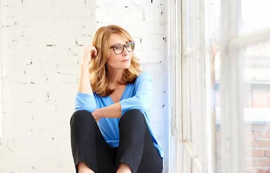 Nachdenklich aussehende mittelalte Frau mit großer Brille sitzt vor einem Fenster und schaut hinaus.