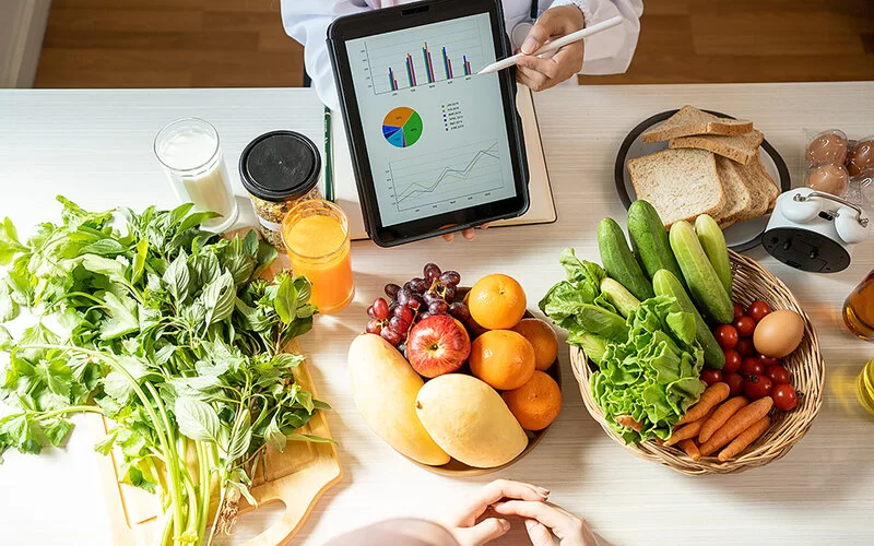 Sicht auf einen Tisch, auf dem verschiedene Lebensmittel stehen: ein Brett mit Kräutern, ein Korb mit Obst, ein Korb mit Gemüse, Toastscheiben, Eier und verschiedene Getränke. An den Längsseiten des Tisches sitzen zwei Personen, deren Köpfe nicht im Bild sind. Eine Person trägt einen weißen Kittel, hält ein Tablet in der Hand und zeigt mit einem Tablet-Stift auf Diagramme auf dem Display.
