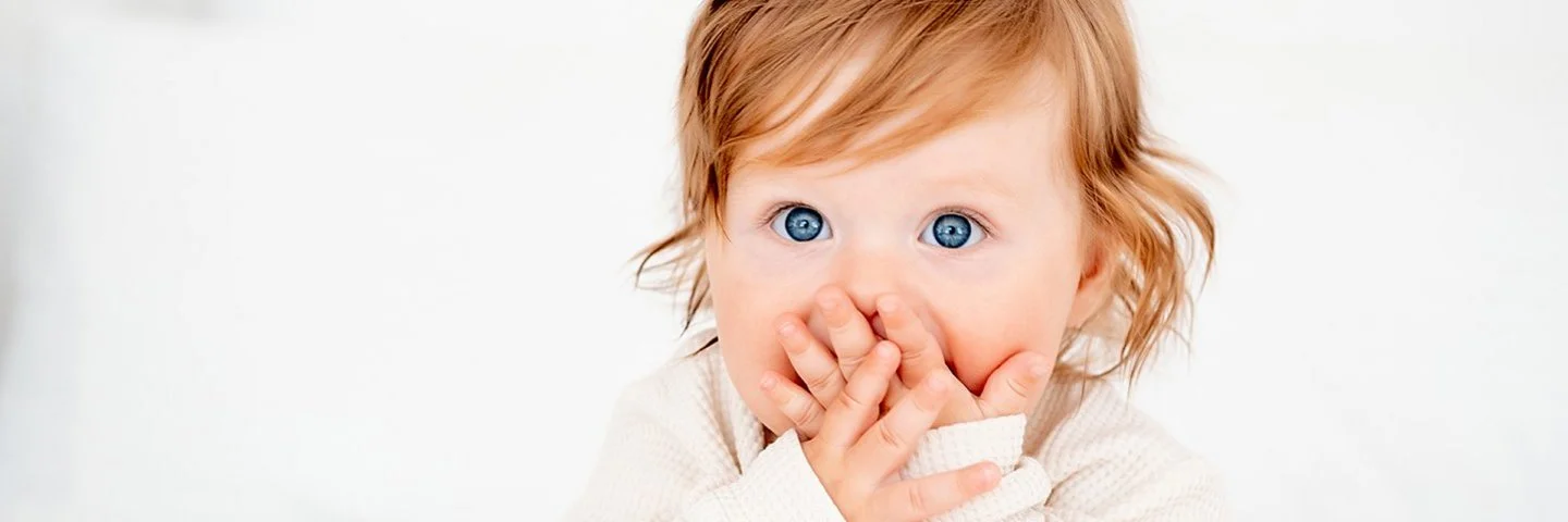 Ein Baby mit roten Haaren und blauen Augen in einem weißen Bodysuit auf einem Bett hält sich erschrocken beide Hände vor den Mund.