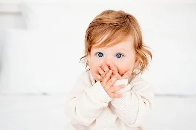 Ein Baby mit roten Haaren und blauen Augen in einem weißen Bodysuit auf einem Bett hält sich erschrocken beide Hände vor den Mund.