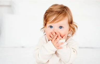 Ein Baby mit roten Haaren und blauen Augen in einem weißen Bodysuit auf einem Bett hält sich erschrocken beide Hände vor den Mund.