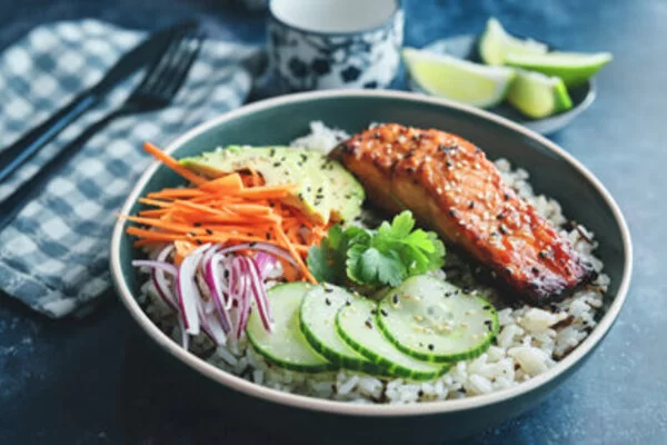 Man sieht eine Bowl mit Reis, Gurken, Zwiebeln, Karotten, Avocado und Lachs.