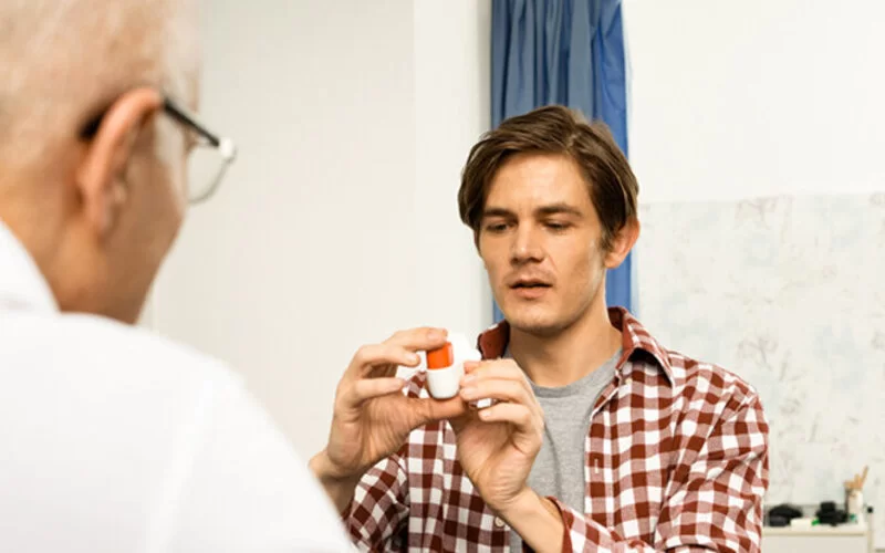 Ein Arzt zeigt einem jungen Mann mit der chronischen Erkrankung Asthma, wie man den Asthmainhalator richtig anwendet.