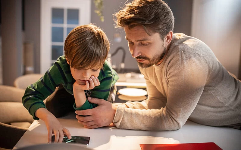 Vater mit Sohn im Grundschulalter lehnen beide an einem Tisch und schauen gemeinsam auf ein Smartphone.