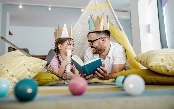 Vater liest seiner Tochter aus einem Buch vor – beide tragen Kronen aus Karton und lächeln sich fröhlich an.