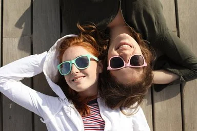 Zwei Frauen liegen nebeneinander auf Holzplanken, eine trägt eine weiße Jacke und gestreiftes Shirt, die andere eine dunkle Jacke und rosa Sonnenbrille