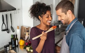 Gesund und günstig kochen: Eine Frau und ein Mann stehen gemeinsam in der Küche und schauen lachend auf einen Kochlöffel mit Tomatensoße.